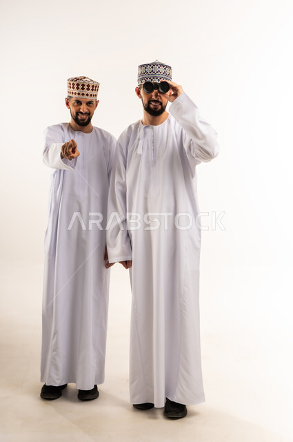 رؤية الأشياء البعيدة, الطموحات والأهداف المستقبلية, مفهوم الاستكشاف والمغامرة, بورتريه لشابين توأم عربيين خليجيين عمانيين يرتديان الدشداشة والكمة أحدهما يشير بإصبع السبابة والآخر يمسك المنظار ثنائي العينين, الجسم بالطول الكامل, خلفية بيضاء