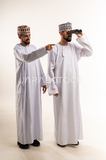 الطموحات والأهداف المستقبلية, مفهوم الاستكشاف والمغامرة, رؤية الأشياء البعيدة, بورتريه لشابين توأم عربيين خليجيين عمانيين يرتديان الدشداشة والكمة أحدهما يشير بإصبع السبابة والآخر يمسك المنظار ثنائي العينين, الجسم بالطول الكامل, خلفية بيضاء