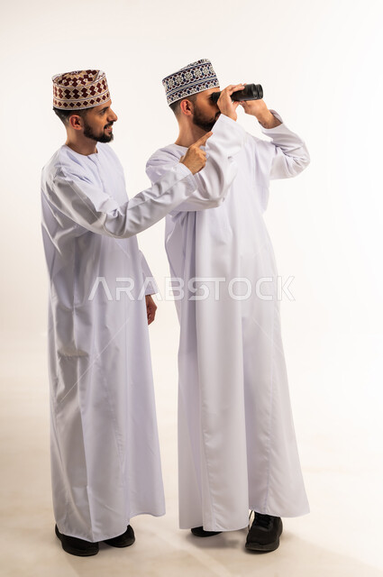 رؤية الأشياء البعيدة, الطموحات والأهداف المستقبلية, مفهوم الاستكشاف والمغامرة, بورتريه لشابين توأم عربيين خليجيين عمانيين يرتديان الدشداشة والكمة أحدهما يشير بإصبع السبابة والآخر يمسك المنظار ثنائي العينين, الجسم بالطول الكامل, خلفية بيضاء