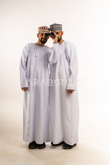 Concept of exploration and adventure, future ambitions and goals, seeing things far away, portrait of two young Omani Gulf Arab twins wearing dishdasha and kummah looking through binoculars, full body, white background