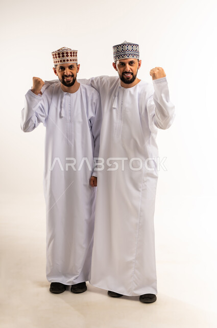 الفوز وتحقيق الأهداف, الفرح لسماع خبر ما, المساندة والدعم بين الأخوة, بورتريه لشابين توأم عربيين خليجيين عمانيين يرتديان الدشداشة والكمة يرفعان قبضة يديهما بتعابير التشجيع والحماس, الجسم بالطول الكامل, خلفية بيضاء