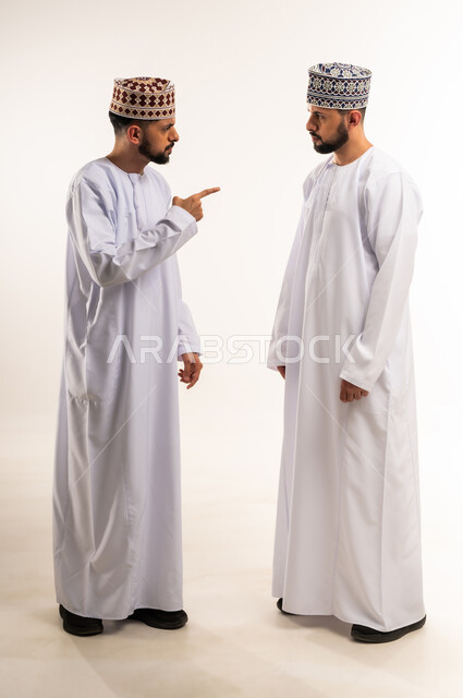 تعابير التوبيخ والغضب, الضغط العصبي و التوتر, النقاش الحاد والمواجهة بين الأخوة, بورتريه لشاب عربي خليجي عماني يرتدي الدشداشة والكمة يقف مع أخيه التوأم ويرفع اصبع السبابة بإيماءات إلقاء اللوم, الجسم بالطول الكامل, خلفية بيضاء