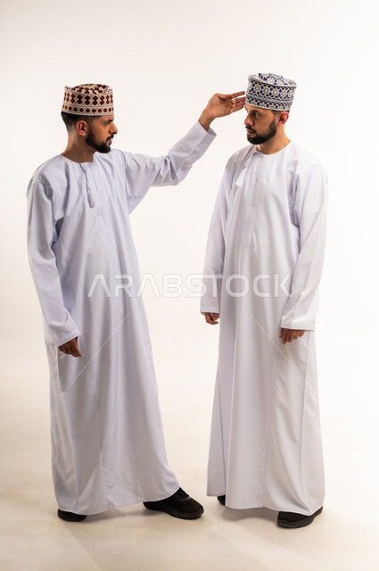 الضغط العصبي و التوتر, تعابير التوبيخ والغضب, النقاش الحاد والمواجهة بين الأخوة, بورتريه لشاب عربي خليجي عماني يرتدي الدشداشة والكمة يقف مع أخيه التوأم ويرفع يده بإيماءات إلقاء اللوم, الجسم بالطول الكامل, خلفية بيضاء