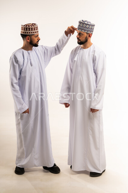 النقاش الحاد والمواجهة بين الأخوة، الضغط العصبي و التوتر, تعابير التوبيخ والغضب, بورتريه لشاب عربي خليجي عماني يرتدي الدشداشة والكمة يقف مع أخيه التوأم ويرفع يده بإيماءات إلقاء اللوم, الجسم بالطول الكامل, خلفية بيضاء