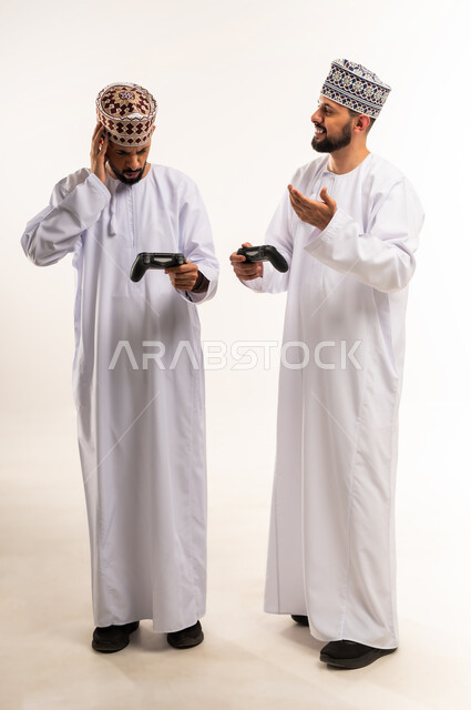 Expressions of sadness for losing the game, joy for winning and triumph, using modern technology for entertainment and amusement, enjoying fun video games, portrait of two young Omani Arab Gulf twins wearing dishdasha and thobe holding joystick in their hands, full body, white background