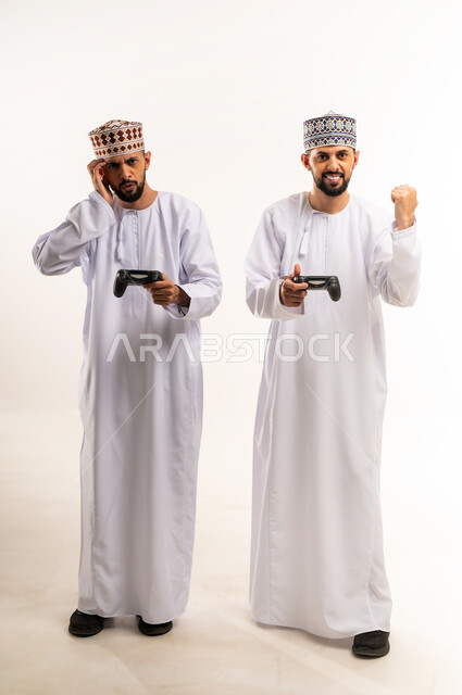 Raising fist in victory gesture, expressions of sadness for loss, using modern technology for entertainment and amusement, enjoying fun video games, portrait of two young Omani Arab Gulf twins wearing dishdasha and kummah holding joysticks in their hands, full body, white background