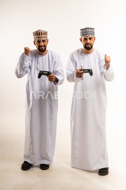 Raising fist with excited expression, having fun with brothers, using modern technology for entertainment, enjoying fun video games, portrait of two young Omani Gulf Arab twins wearing dishdasha and kummah holding joystick in their hands, full body, white background