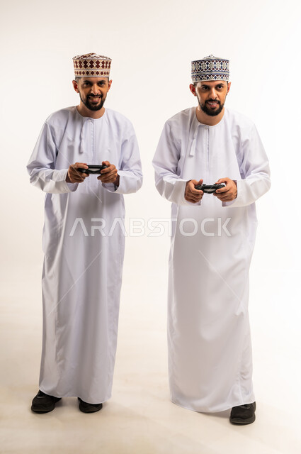 Using modern technology for entertainment and fun, spending quality time with brothers, enjoying fun video games, portrait of two young Omani Gulf Arab twins wearing dishdasha and thobe holding joysticks in their hands, full body, white background
