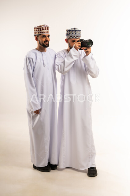 استخدام تقنية التصوير الحديثة والمبتكرة, الشغف بممارسة الهواية المفضلة, توثيق اللحظات السعيدة, بورتريه لشابين توأم عربيين خليجيين عمانيين يرتديان الدشداشة والكمة يستخدمان كاميرا فوتوغرافية عالية الدقة, الجسم بالطول الكامل, خلفية بيضاء