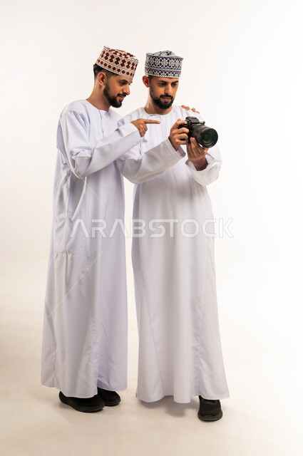 Passion for practicing a favorite hobby, documenting happy moments, using modern and innovative photography technology, portrait of two young Arab Gulf Omani twins wearing a dishdasha and a kummah using a high-resolution photographic camera, full-length body, white background