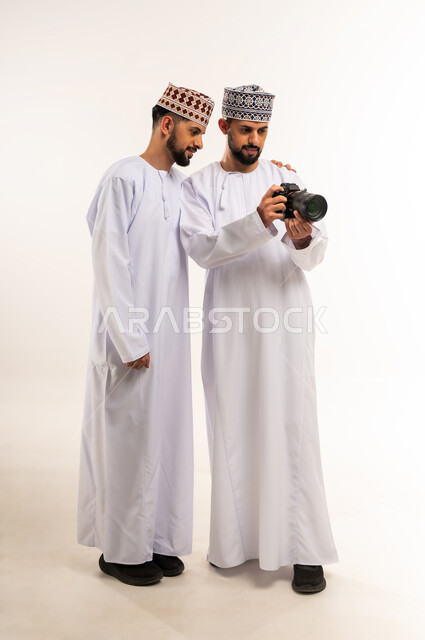 Using modern and innovative photography technology, passion for practicing a favorite hobby, documenting happy moments, portrait of two young Arab Gulf Omani twins wearing a dishdasha and a kummah using a high-resolution photographic camera, full-length body, white background