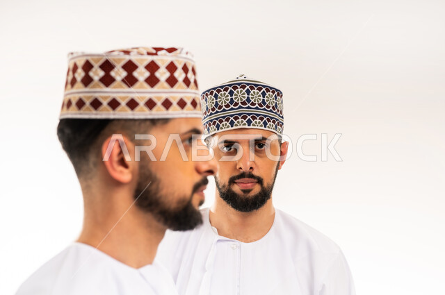 الاهتمام بالأناقة والمظهر الخارجي, إيماءات حدة النظر والتحديق إلى الكاميرا,، بورتريه مقرب لشابين توأم عربيين خليجيين عمانيين يرتديان الدشداشة والكمة أحدهما ينظر إلى الجانب والآخر ينظر إلى الكاميرا، خلفية بيضاء