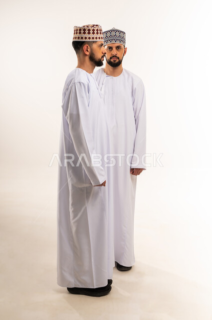 إيماءات حدة النظر والتحديق إلى الكاميرا, الاهتمام بالأناقة والمظهر الخارجي، بورتريه لشابين توأم عربيين خليجيين عمانيين يرتديان الدشداشة والكمة أحدهما ينظر إلى الجانب والآخر ينظر إلى الكاميرا, الجسم بالطول الكامل، خلفية بيضاء