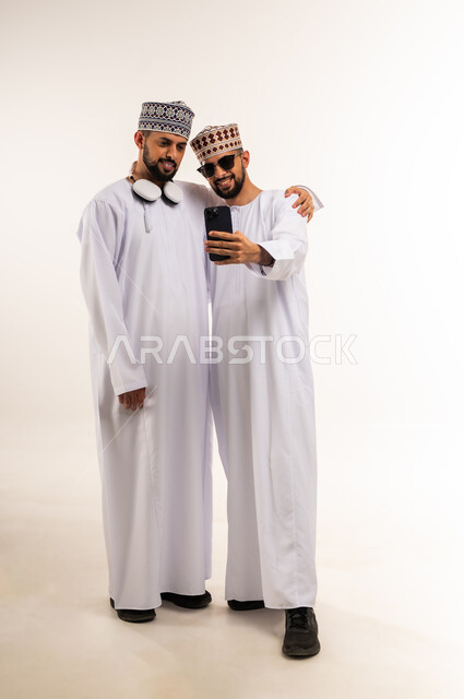 توثيق اللحظات السعيدة عبر الهاتف المحمول, استخدام جهاز تقني حديث ومتطور, استعمال سماعات الرأس الصوتية, بورتريه لشاب عربي خليجي عماني يرتدي الدشداشة والكمة يقوم بالتقاط صورة السيلفي التذكارية مع أخيه التوأم عبر الجوال, الجسم بالطول الكامل, خلفية بيضاء
