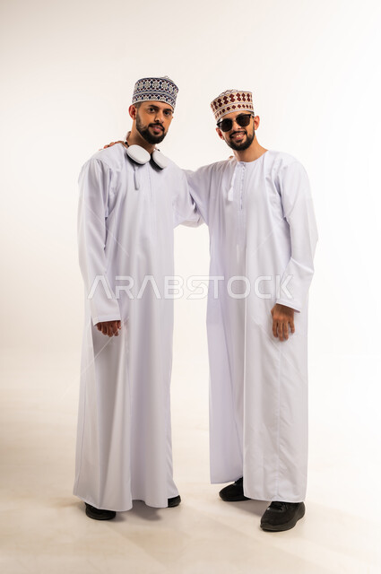 قضاء الوقت الممتع مع الأخوة, الاستمتاع بسماع الموسيقا المفضلة, مشاركة الأشقاء اللحظات السعيدة, بورتريه لشابين توأم عربيين خليجيين عمانيين يرتديان الدشداشة والكمة أحدهما يضع نظارة شمسية والآخر سماعات الرأس الصوتية, الجسم بالطول الكامل, خلفية بيضاء