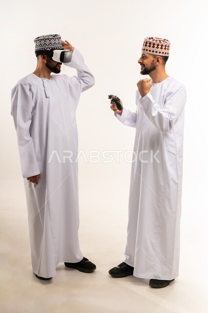 رفع قبضة اليد بإيماءات الفوز والحماس, الاستمتاع بألعاب الفيديو الممتعة, محاكاة العالم الخيالي, بورتريه لشابين توأم عربيين خليجيين عمانيين يرتديان الدشداشة والكمة إحداهما يمسك عصا التحكم والآخر يضع نظارة الفي ار, الجسم بالطول الكامل, خلفية بيضاء