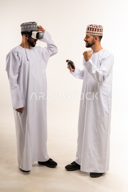 الاستمتاع بألعاب الفيديو الممتعة, رفع قبضة اليد بإيماءات الفوز والحماس, محاكاة العالم الخيالي, بورتريه لشابين توأم عربيين خليجيين عمانيين يرتديان الدشداشة والكمة إحداهما يمسك عصا التحكم والآخر يضع نظارة الفي ار, الجسم بالطول الكامل, خلفية بيضاء