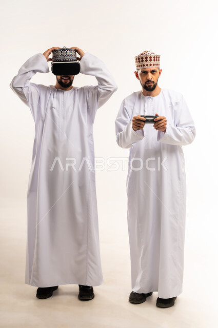 استخدام التكنولوجيا الحديثة للتسلية والترفيه, محاكاة العالم الخيالي, الاستمتاع بألعاب الفيديو الممتعة, بورتريه لشابين توأم عربيين خليجيين عمانيين يرتديان الدشداشة والكمة إحداهما يمسك عصا التحكم والآخر يضع نظارة الفي ار, الجسم بالطول الكامل, خلفية بيضاء