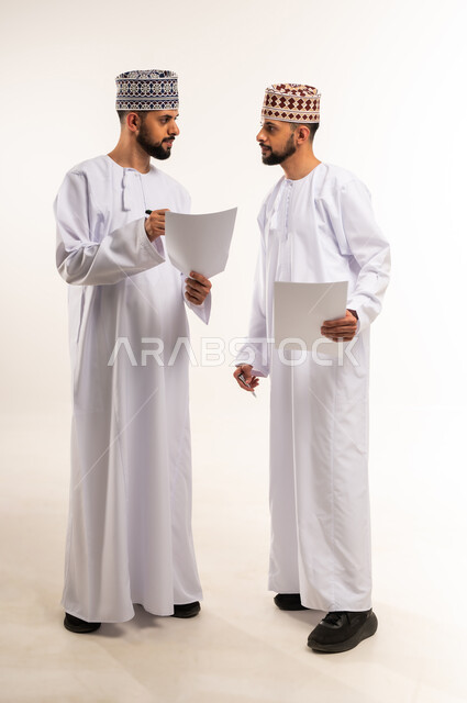 Cooperation between brothers in completing work tasks, exchanging experiences and skills, discussing plans and projects, portrait of two young Omani Gulf Arab twins wearing a dishdasha and a kummah holding white papers in their hands, full-length body, white background