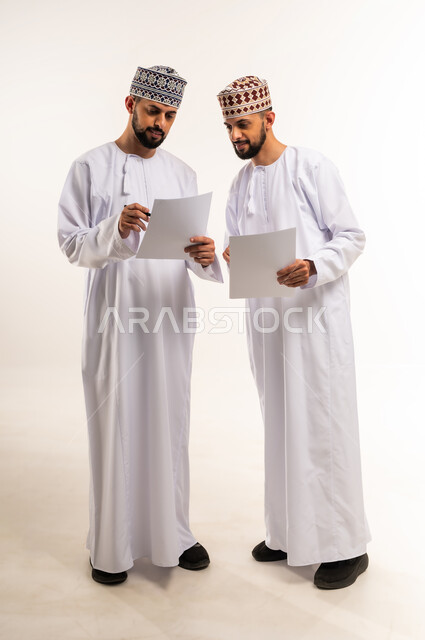 التعاون بين الأخوة في إنجاز مهام العمل, تبادل الخبرات والمهارات, مناقشة الخطط والمشاريع, بورتريه لشابين توأم عربيين خليجيين عمانيين يرتديان الدشداشة والكمة يمسكان بأيديهم الأوراق البيضاء, الجسم بالطول الكامل, خلفية بيضاء