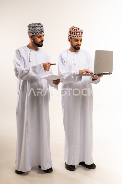 تبادل الخبرات والمهارات, مناقشة الخطط والمشاريع, التعاون بين الأخوة في إنجاز مهام العمل, استخدام جهاز تقني حديث ومتطور, بورتريه لشابين توأم عربيين خليجيين عمانيين يرتديان الدشداشة والكمة أحدهما يمسك الحاسوب المحمول والأخر الأوراق, الجسم بالطول الكامل, خلفية بيضاء