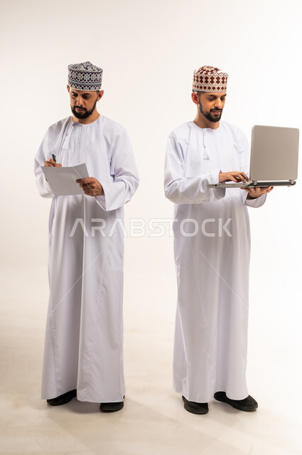 مناقشة الخطط والمشاريع, تبادل الخبرات والمهارات, التعاون بين الأخوة في إنجاز مهام العمل, استخدام جهاز تقني حديث ومتطور, بورتريه لشابين توأم عربيين خليجيين عمانيين يرتديان الدشداشة والكمة أحدهما يمسك الحاسوب المحمول والآخر الأوراق, الجسم بالطول الكامل, خلفية بيضاء