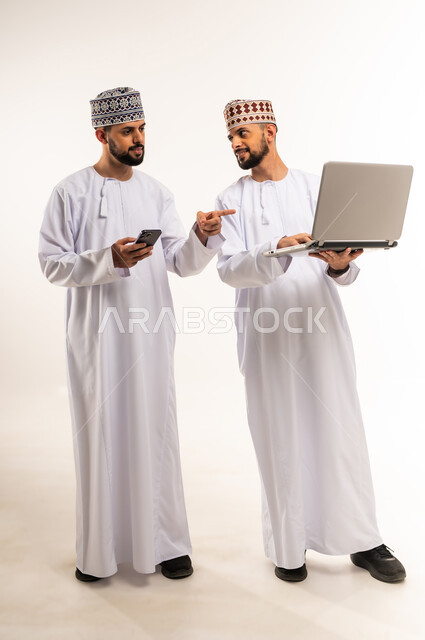 Completing work tasks remotely, using modern and advanced technical devices, portrait of a young Arab Gulf Omani man wearing a dishdasha and a kummah standing with his twin brother holding a mobile phone in his hand and pointing his index finger at the laptop, full-length body, white background