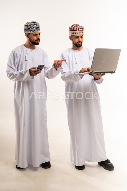استخدام الأجهزة التقنية الحديثة والمتطورة, إنجاز مهام العمل عن بعد, بورتريه لشاب عربي خليجي عماني يرتدي الدشداشة والكمة يقف مع أخيه التوأم يمسك بيده الجوال ويشير بإصبع السبابة إلى الحاسوب المحمول, الجسم بالطول الكامل, خلفية بيضاء