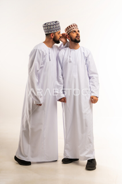 التكلم بصوت منخفض, النميمة والتحدث عن شخص ما، الانصات والتركيز بالكلام، مفهوم الكتمان وحفظ الأسرار بين الأشقاء, بورتريه لشاب عربي خليجي عماني يرتدي الدشداشة والكمة يهمس بإذن أخيه التوأم, الجسم بالطول الكامل, خلفية بيضاء