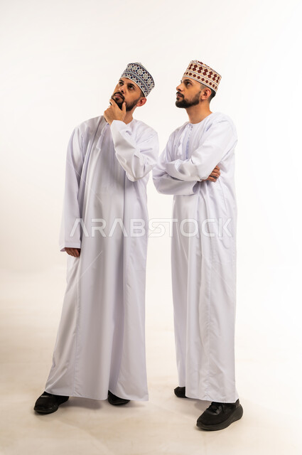 التأمل و التخطيط للمستقبل, إيماءات شرود الذهن والحيرة, محاولة إيجاد حل لمشكلة ما, بورتريه لشابين توأم عربيين خليجيين عمانيين يرتديان الدشداشة والكمة ينظران للأعلى ويضعان يدهما على ذقنهما بإيماءات التمعن والتركيز, الجسم بالطول الكامل, خلفية بيضاء