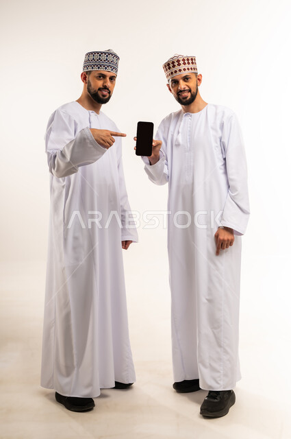 الإشارة بإصبع السبابة للهاتف المحمول, التسلية والترفيه في وقت الفراغ, استخدام جهاز تقني حديث ومتطور, بورتريه لشاب عربي خليجي عماني يرتدي الدشداشة والكمة يقف مع أخيه التوأم ويمسك بيده الجوال, عرض شاشة سوداء فارغة, الجسم بالطول الكامل, خلفية بيضاء