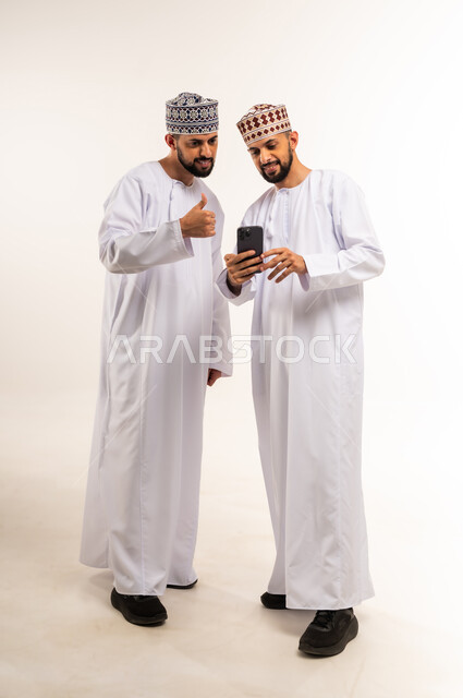 الإشارة بإصبع الإبهام للأعلى, استخدام جهاز تقني حديث ومتطور, تعابير الموافقة والقبول, إيماءات الرضا والجودة والامتياز, بورتريه لشابين عربيين خليجيين عمانيين يرتديان الدشداشة والكمة يقومان بإجراء مكالمة فيديو عبر الهاتف المحمول, الجسم بالطول الكامل, خلفية بيضاء
