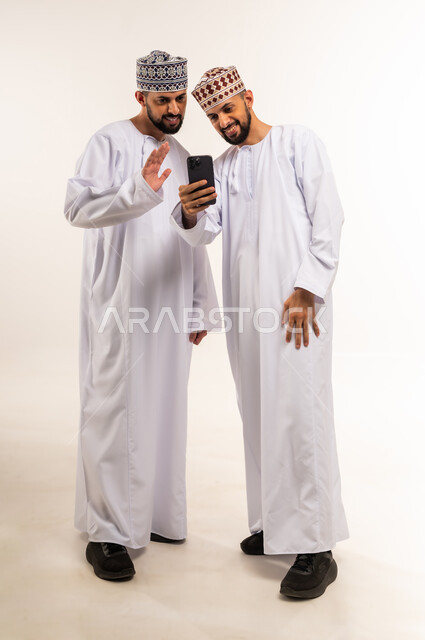 تعابير إلقاء السلام والتحية, استخدام جهاز تقني حديث ومتطور, التواصل مع الأصدقاء والعائلة, بورتريه لشابين توأم عربيين خليجيين عمانيين يرتديان الدشداشة والكمة يقومان بإجراء مكالمة فيديو عبر الهاتف المحمول, الجسم بالطول الكامل, خلفية بيضاء