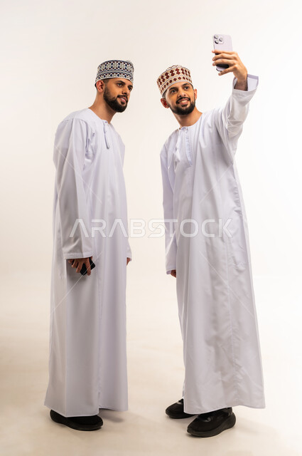 توثيق اللحظات السعيدة عبر الهاتف المحمول, استخدام جهاز تقني حديث ومتطور, قضاء الوقت الممتع مع الاخوة, بورتريه لشاب عربي خليجي عماني يرتدي الدشداشة والكمة يقوم بالتقاط صورة السيلفي التذكارية مع أخيه التوأم عبر الجوال, الجسم بالطول الكامل, خلفية بيضاء