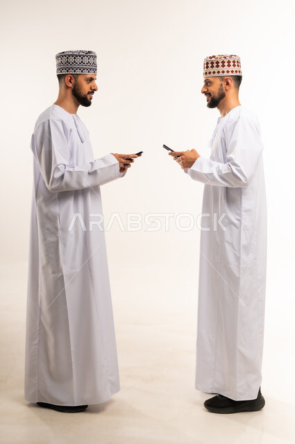 تصفح وسائل التواصل الاجتماعي, التسلية والترفيه في وقت الفراغ, استخدام جهاز تقني حديث ومتطور, بورتريه من الجانب لشابين توأم عربيين خليجيين عمانيين يرتديان الدشداشة والكمة يقومان باستعمال الهاتف المحمول, الجسم بالطول الكامل, خلفية بيضاء