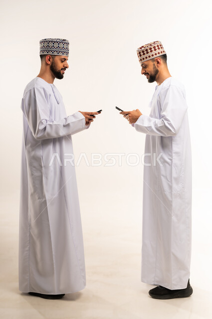 Leisure and entertainment, browsing social media, using a modern and advanced technical device, side portrait of two young Omani Arab Gulf twins wearing a dishdasha and a kimono using a mobile phone, full body, white background