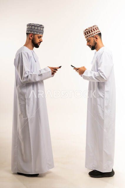 Using a modern and advanced technical device, entertainment and leisure time, browsing social media, side portrait of two young Omani Arab Gulf twins wearing a dishdasha and a kimono using a mobile phone, full body, white background
