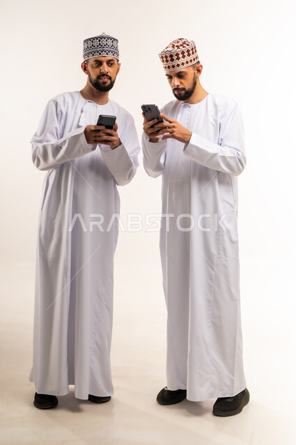 Snooping and sneaking glances at another's mobile phone, using a modern and advanced technical device, browsing social media, portrait of two young Omani Arab Gulf twins wearing a dishdasha and a thobe using a mobile phone, full body, white background