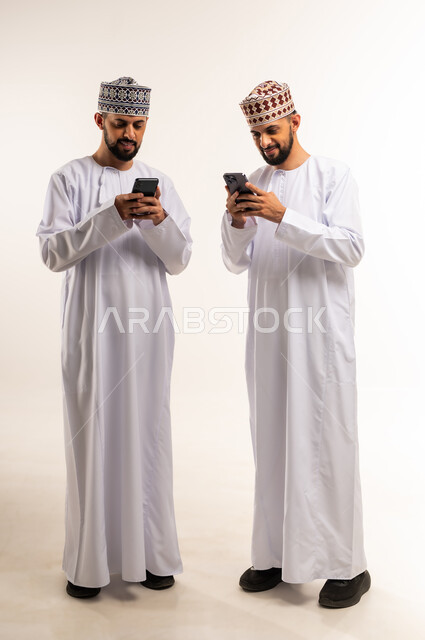 Entertainment and leisure time, using a modern and advanced technical device, browsing social media, portrait of two young Omani Arab Gulf twins wearing a dishdasha and a keffiyeh using a mobile phone, full body, white background