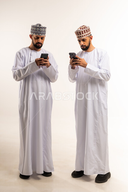 استخدام جهاز تقني حديث ومتطور, التسلية والترفيه في وقت الفراغ, تصفح وسائل التواصل الاجتماعي, بورتريه لشابين توأم عربيين خليجيين عمانيين يرتديان الدشداشة والكمة يقومان باستعمال الهاتف المحمول, الجسم بالطول الكامل, خلفية بيضاء