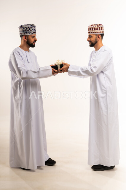 تقديم الهدايا بالأعياد والمناسبات السعيدة, التعبير عن المحبة والتقدير, الاحتفال بعيد الميلاد, بورتريه لشاب عربي خليجي عماني يرتدي الدشداشة والكمة يقوم بإهداء أخاه التوأم علبة هدية, الجسم بالطول الكامل, خلفية بيضاء