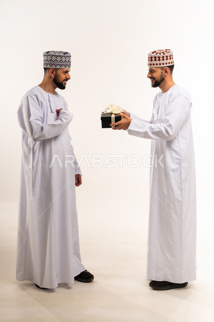 التعبير عن المحبة والتقدير, تقديم الهدايا بالأعياد والمناسبات السعيدة, الاحتفال بعيد الميلاد, بورتريه لشاب عربي خليجي عماني يرتدي الدشداشة والكمة يقوم بإهداء أخاه التوأم علبة هدية, الجسم بالطول الكامل, خلفية بيضاء