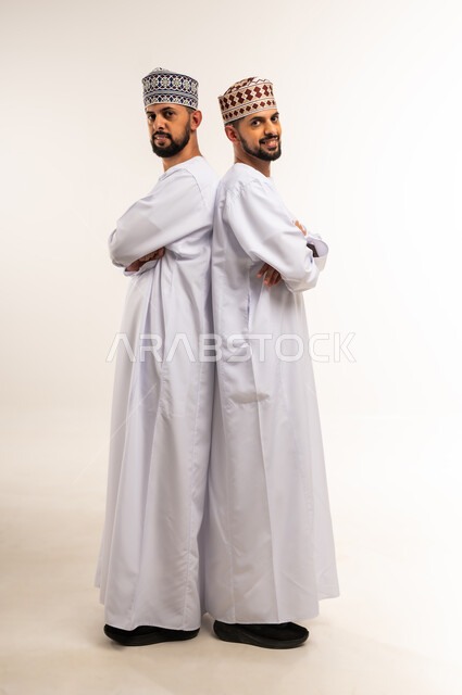Concept of elegance and masculinity, concern for outward appearance, looking at the camera with gestures of happiness and pleasure, side portrait of two young Omani Arab Gulf twins wearing dishdasha and kummah standing straight with crossed hands, full body, white background