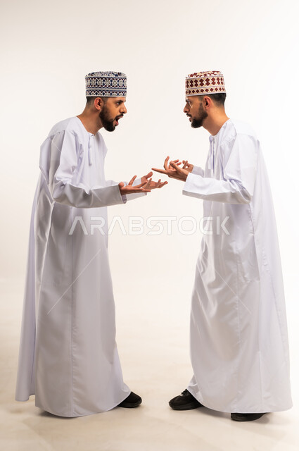 التعصب والتوتر الانفعالي, النقاش والمواجهة بين الاخوات, سوء التفاهم بين الاخوة، بورتريه من الجانب لشابين توأم عربيين خليجيين عمانيين يرتديان الدشداشة والكمة يقفان مقابل بعضهما ويتجادلان بإيماءات الغضب، الجسم بالطول الكامل، خلفية بيضاء