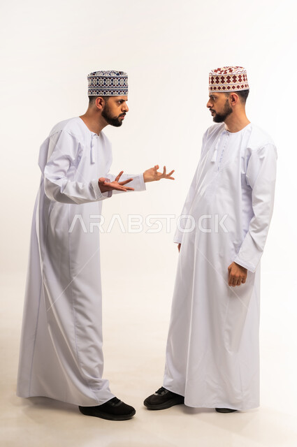 سوء التفاهم بين الاخوة ،النقاش والمواجهة بين الاخوات، بورتريه من الجانب لشابين توأم عربيين خليجيين عمانيين يرتديان الدشداشة والكمة يقفان مقابل بعضهما ويتجادلان بإيماءات الغضب، التعصب والتوتر الانفعالي، الجسم بالطول الكامل، خلفية بيضاء