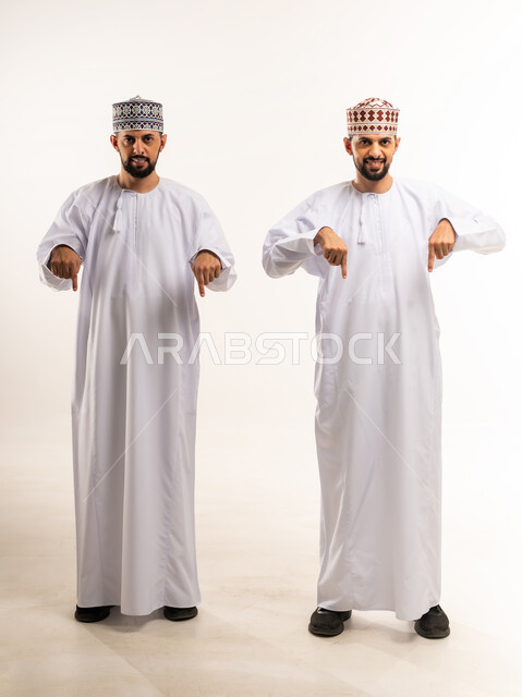 تأكيد حدوث شيء هذه اللحظة, حركات تدل على هذا المكان (هنا)، تحديد موقع معين, بورتريه لشابين توأم عربيين خليجيين عمانيين يرتديان الدشداشة والكمة يشيران بإصبع السبابة للأسفل, الجسم بالطول الكامل, خلفية بيضاء