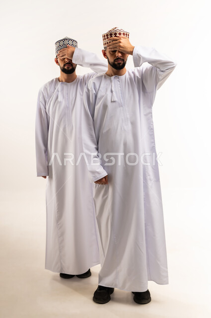 إيماءات وضع الكف على الوجه, حركات تدل على اللعب و المرح, تعابير الخجل و الاستعداد لرؤية المفاجأة, بورتريه لشابين توأم عربيين خليجيين عمانيين يرتديان الدشداشة والكمة يغمضان عينيهما بأيديهما, الجسم بالطول الكامل، خلفية بيضاء