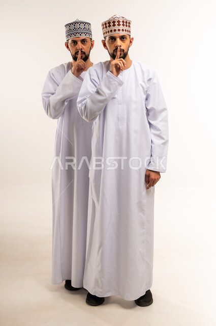الصمت وعدم السماح بالتكلم, النظر إلى الكاميرا بتعابير طلب الهدوء وعدم الازعاج ،السكوت و التوقف عن الكلام, بورتريه لشابين توأم عربيين خليجيين عمانيين يرتديان الدشداشة والكمة يضعان اصبع الإبهام على فمهما, الجسم بالطول الكامل, خلفية بيضاء