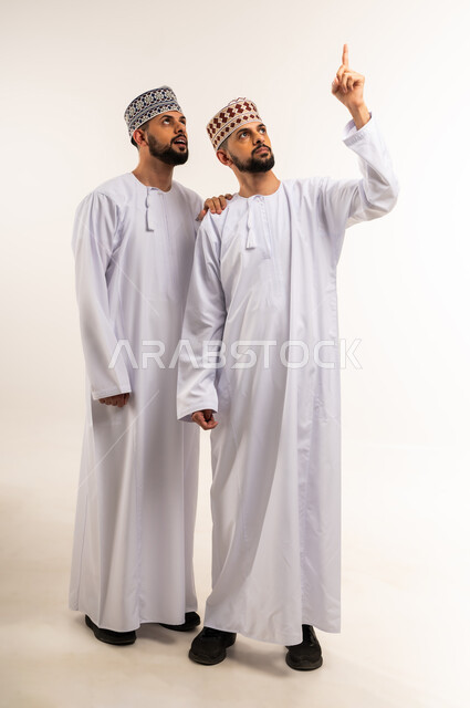 الدلالة على موقع معين, حركات تدل على هذا المكان (هنا), تحديد مكان عنصر محدد, بورتريه لشاب عربي خليجي عماني يرتدي الدشداشة والكمة يشير لأخيه التوأم بإصبع السبابة إلى شيء ما بالأعلى, الجسم بالطول الكامل, خلفية بيضاء