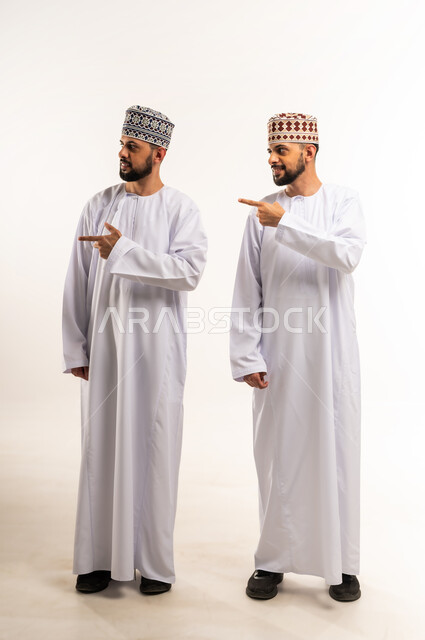 Gestures indicating this place (here), indicating a specific location, locating a specific item, Portrait of two young Omani Gulf Arab twins wearing a dishdasha and a thobe pointing with their index fingers at something, full body, white background